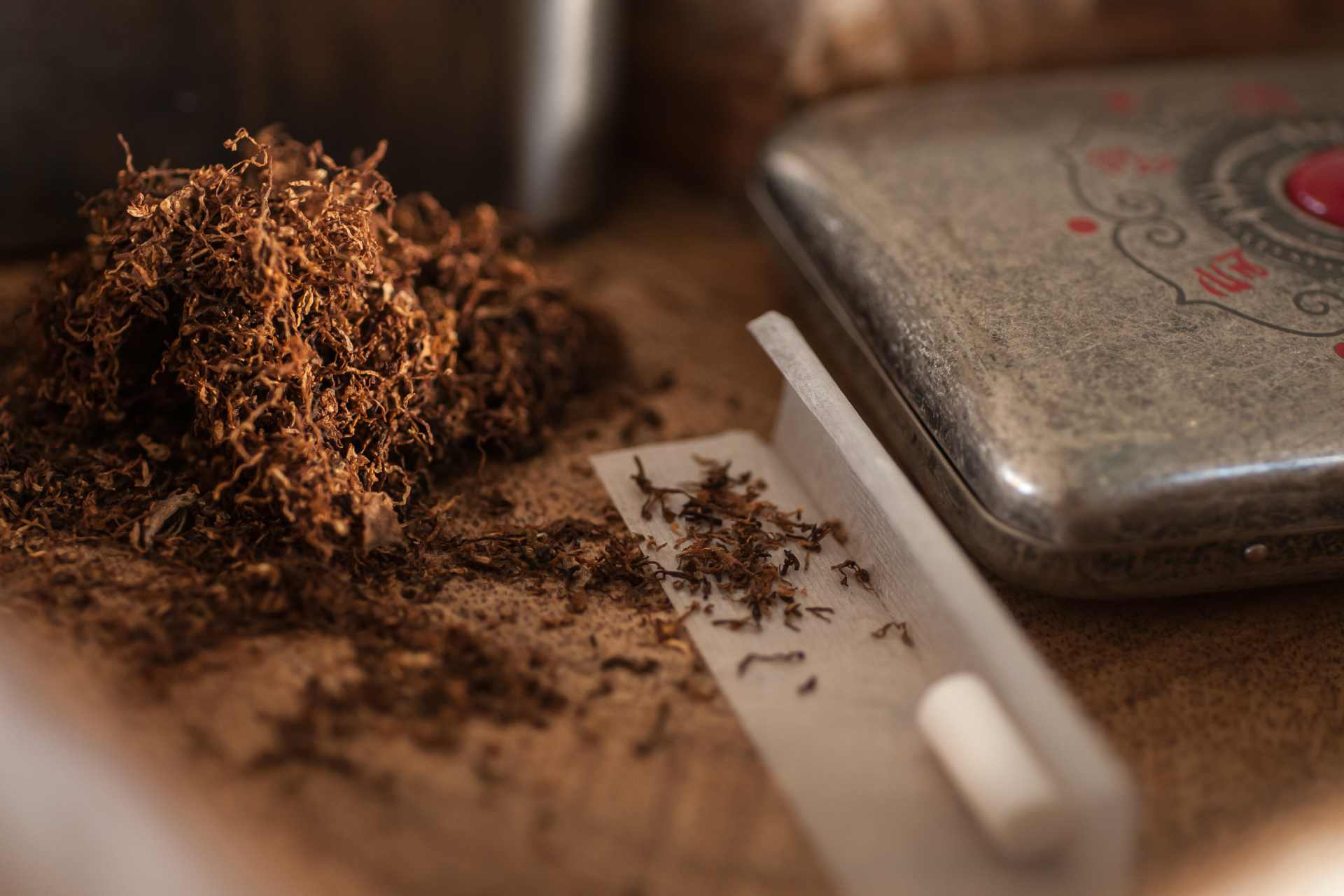 Close up of smoking tobacco on a table beside paper and a filter with an old tobacco tin in the background.