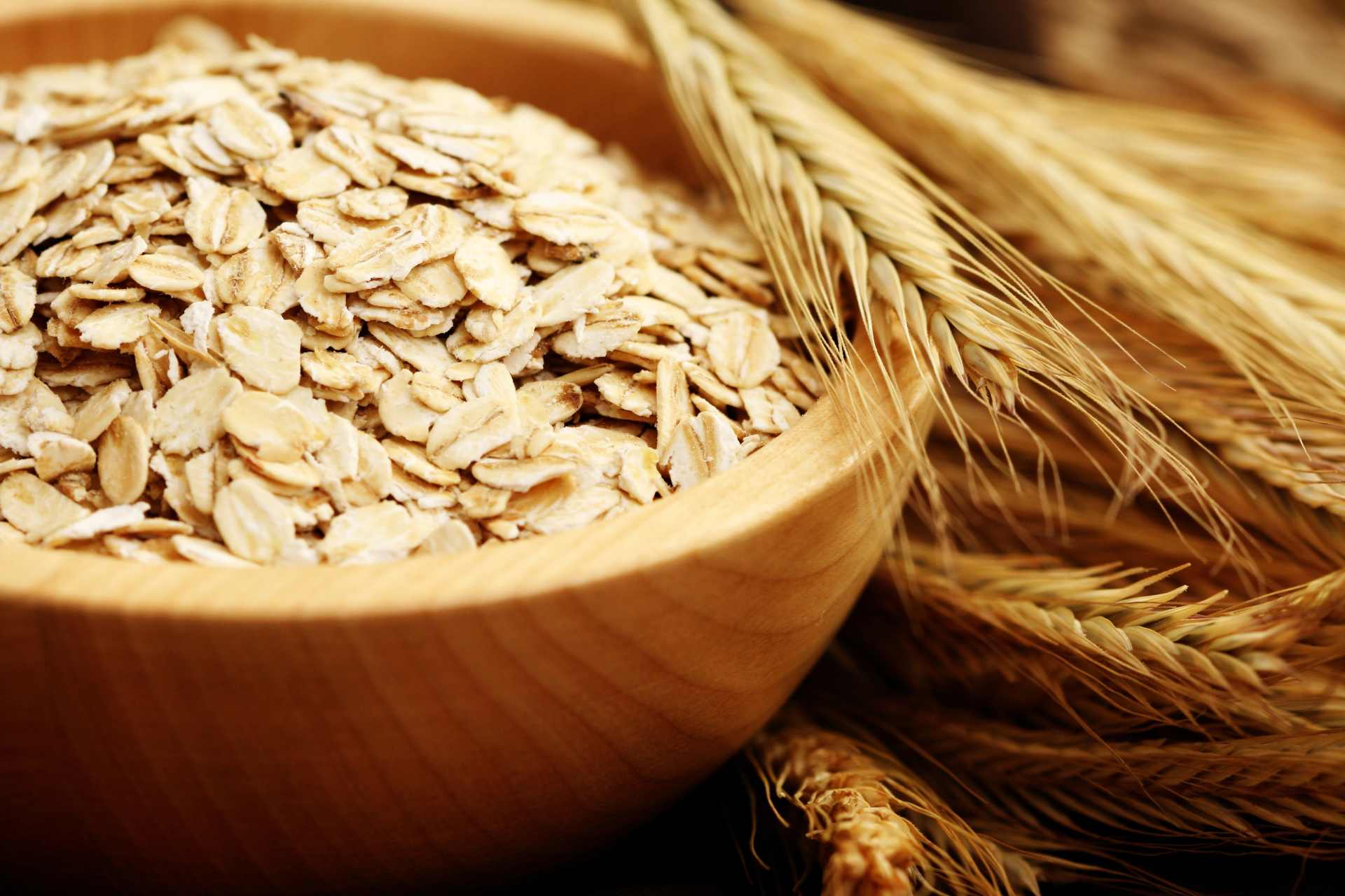 A wooden bowl containing raw oats beside a bunch of oat straw.