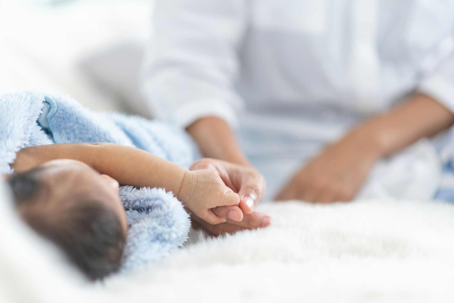Close up of a baby lying on a blue blanket with his mother holding his hand.