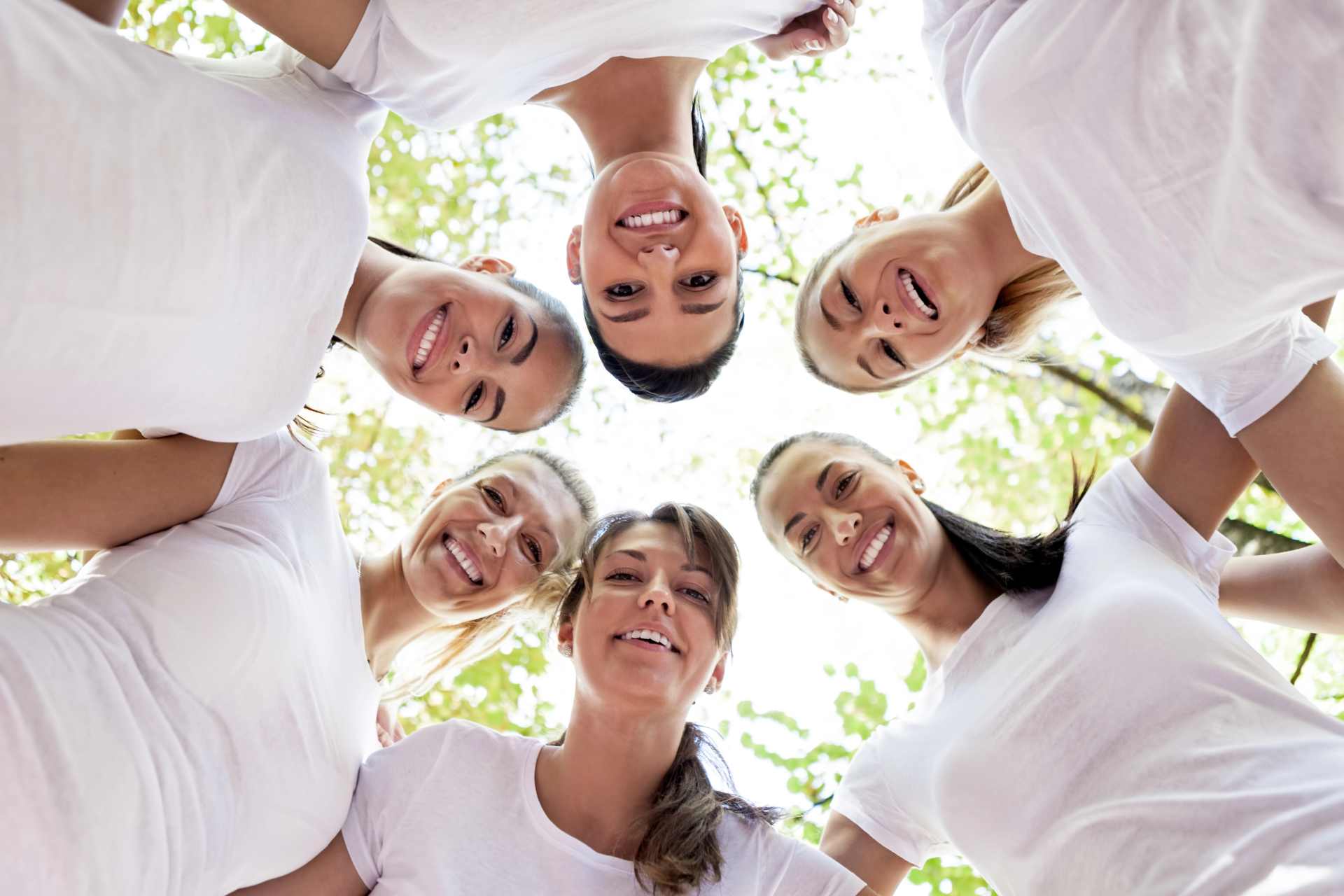 Six women standing in a circle looking down toward a camera placed on the ground, with a blue sky above, symbolizing solidarity, shared vulnerability, and women’s health risks from glyphosate exposure.