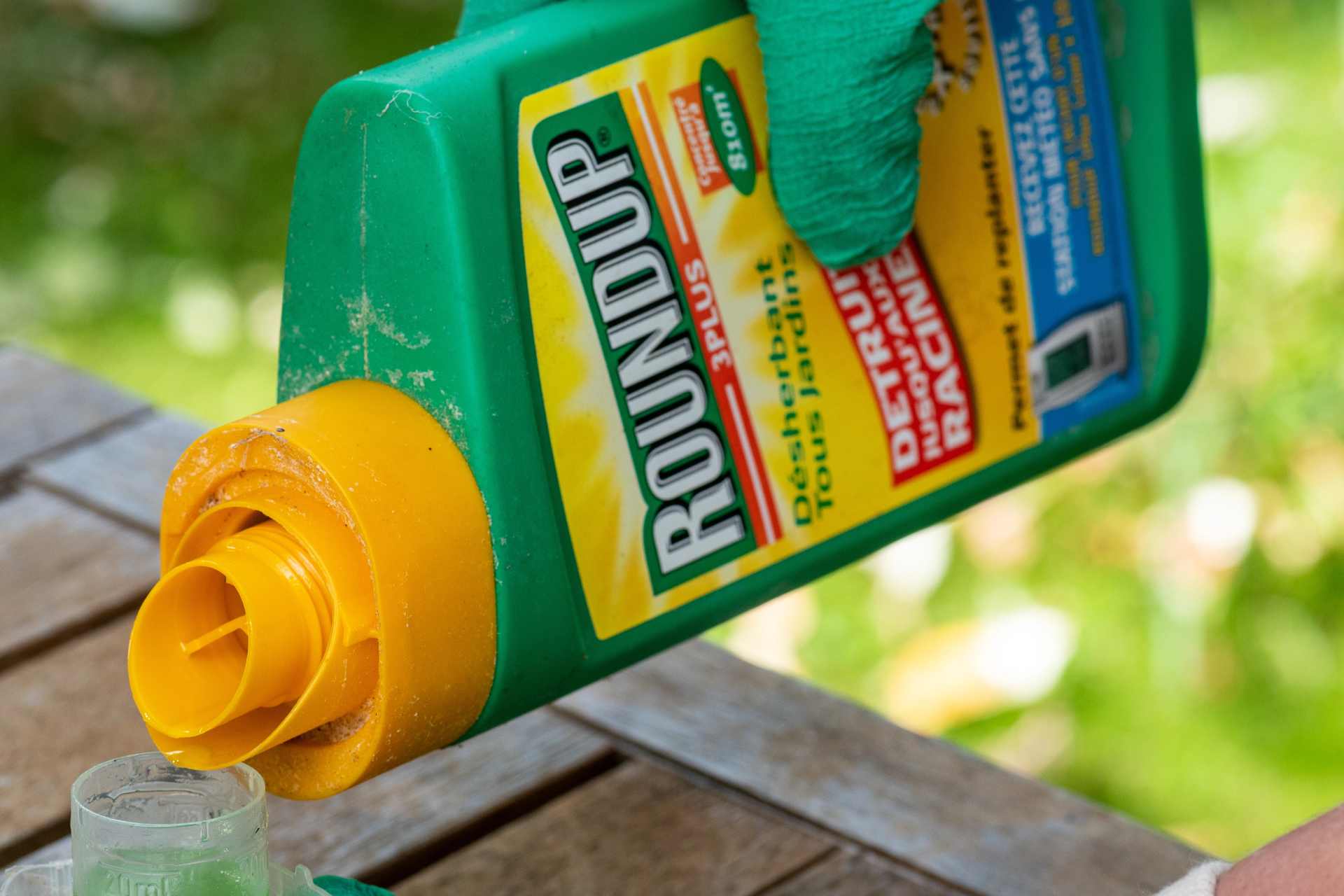 Close-up of Roundup weedkiller being poured into a plastic measuring cup on an outdoor wooden table, highlighting everyday use of glyphosate-based products.