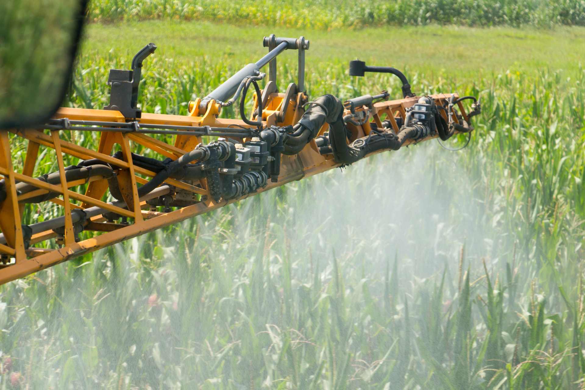 Close-up of a tractor's spray boom releasing herbicide across a green crop field, illustrating the widespread agricultural application of chemical weedkillers like Icafolin and glyphosate.