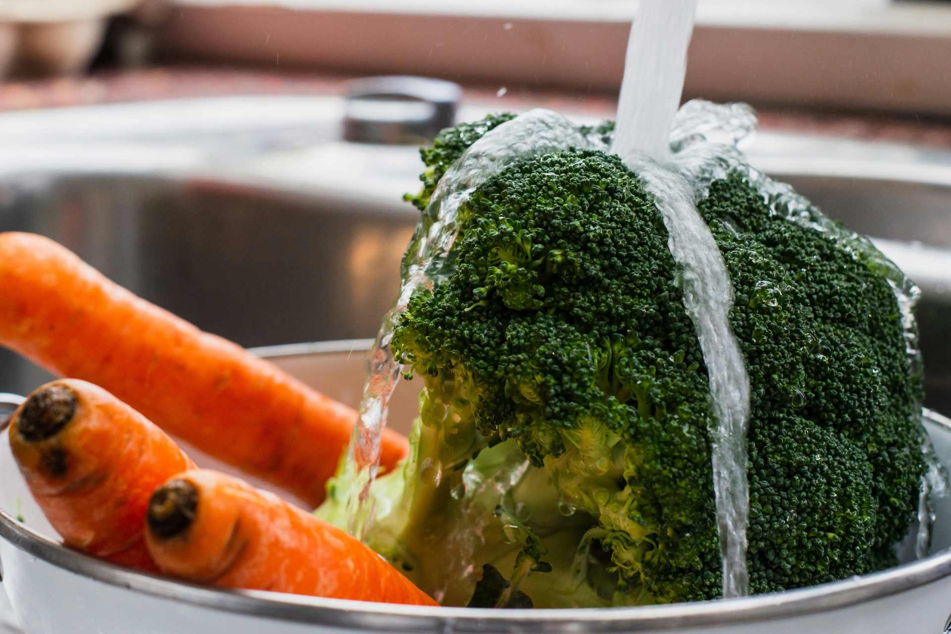 Fresh broccoli and carrots being rinsed under running water in a bowl, illustrating common produce washing practices.