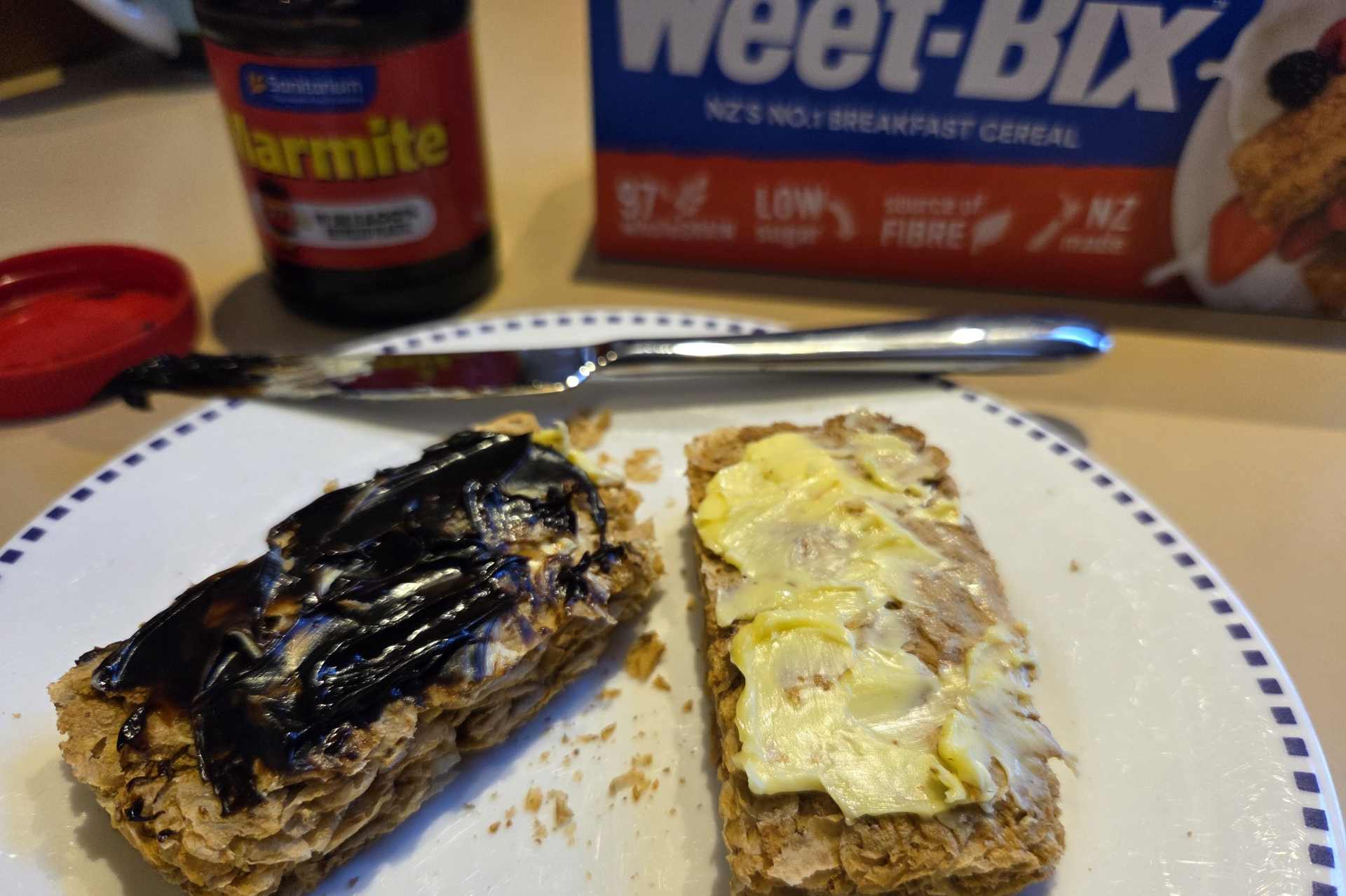 Two Weet-Bix topped with butter and Marmite on a plate, with a jar of Marmite and a packet of Weet-Bix in the background.