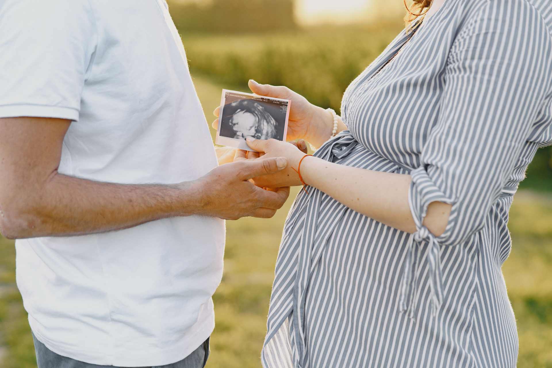 A pregnant woman and a man stand together, both looking down at an ultrasound photo held between them.