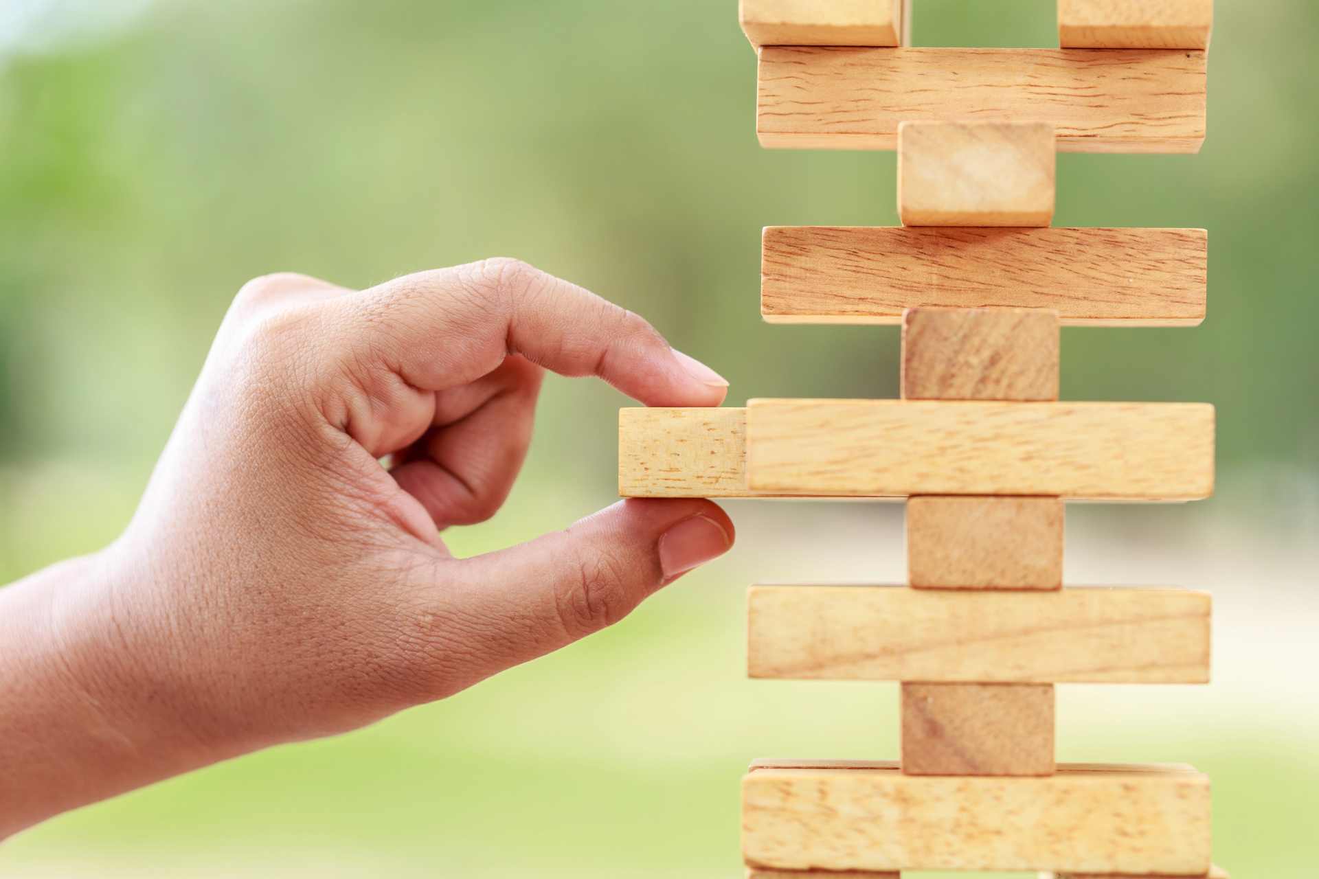 A hand carefully removing a wooden block from a Jenga tower, illustrating the cumulative factors that may contribute to health outcomes and the uncertainty faced by families after a serious illness.